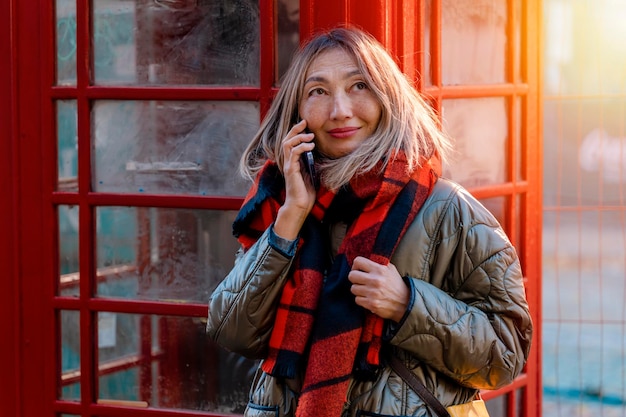 Mujer asiática visitando el Reino Unido usando un teléfono móvil y haciendo un selfie cerca de una cabina de teléfono roja en una ciudad
