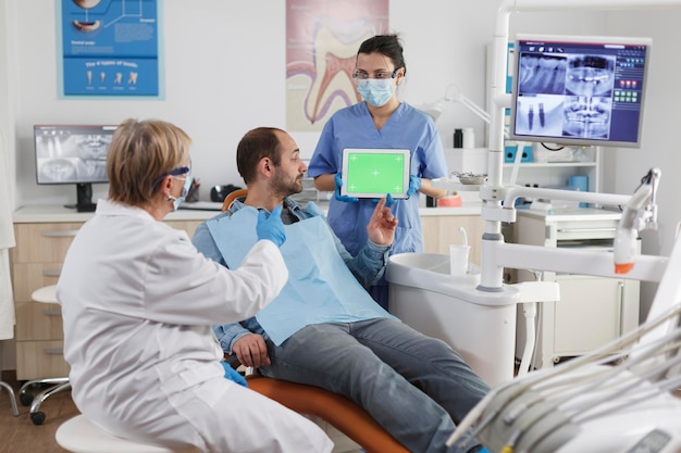 Mujer asistente sosteniendo una tableta de clave de croma de pantalla verde simulada con una pantalla aislada discutiendo la higiene bucal con el paciente durante la consulta estomatológica en el consultorio dental. concepto de estomatología