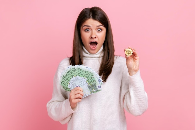 Mujer asombrada con el pelo oscuro que muestra un gran admirador de los billetes en euros y la inversión en bitcoins de oro con un suéter blanco de estilo casual Foto de estudio interior aislada en un fondo rosa