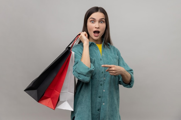 Mujer asombrada de pie con la boca abierta apuntando a bolsas de compras en sus manos sorprendida