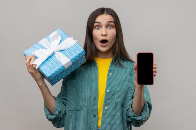 Mujer asombrada sosteniendo caja de regalo y teléfono celular con pantalla vacía para publicidad de compras en línea