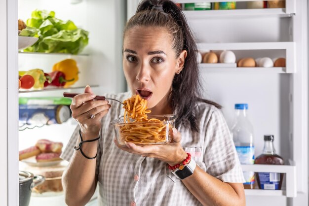 Una mujer asustada y hambrienta en pijama está comiendo espagueti en el refrigerador por la noche.