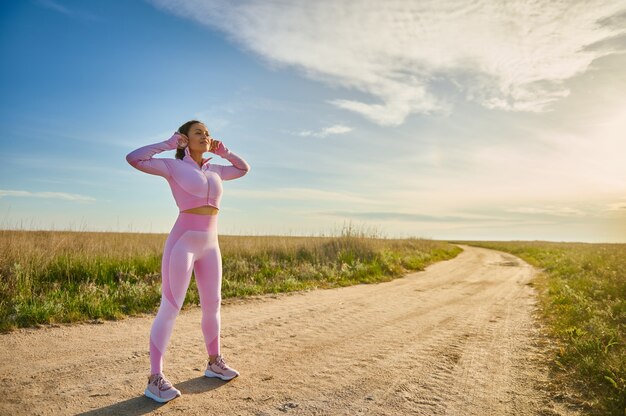 Mujer atleta de cuerpo entero en auriculares y elegante ropa deportiva rosa de pie en la carretera de la estepa lista para trotar en la naturaleza a la luz del atardecer