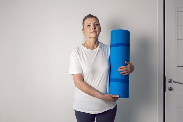 Mujer atleta de edad se encuentra en una habitación blanca con una estera de yoga