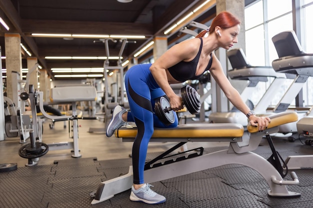 Mujer atleta en ropa deportiva entrenando con pesas en una mano apoyada en el banco en el gimnasio