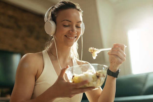 Mujer atlética feliz comiendo ensalada mientras escucha música con auriculares y se relaja en casa