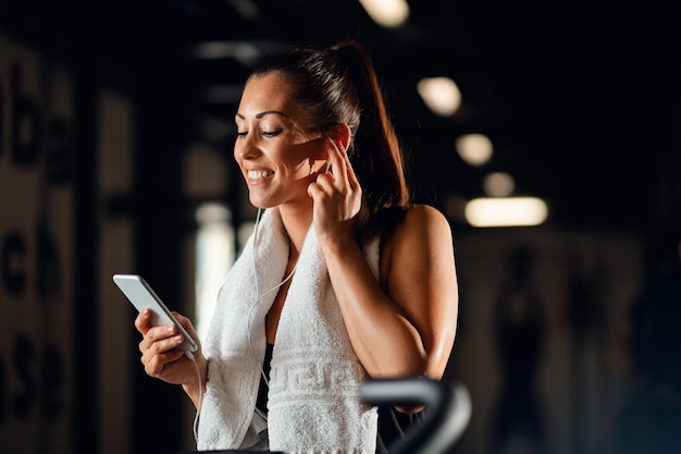 Mujer atlética feliz tomando un descanso después de hacer ejercicio y escuchando música mientras usa un teléfono inteligente en el gimnasio