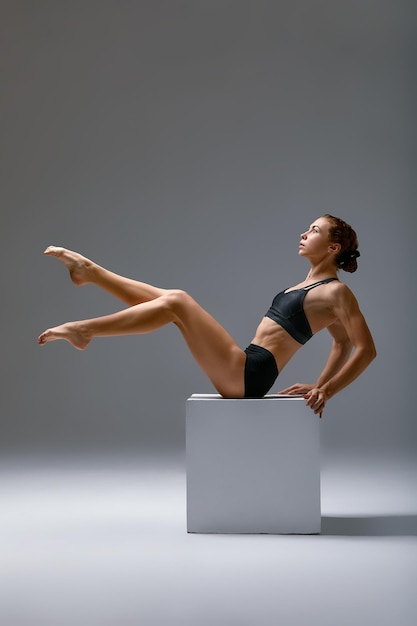 Mujer atlética flexible en ropa deportiva haciendo divisiones en un estudio de fitness sobre un fondo gris Ejercicios de estiramiento con un cubo