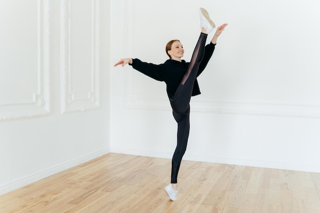 Una mujer atlética en forma demuestra equilibrios de resiliencia en una pierna siendo una gimnasta deportiva experta vestida con ropa deportiva y zapatillas de deporte tiene soportes de ejercicios de pilates en una sala vacía tiene una figura hermosa