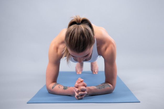 Mujer atlética joven haciendo tablón Mujer fitness de cuerpo entero en ropa deportiva blanca sentada en la alfombra del gimnasio y practicando yoga aislado sobre fondo gris