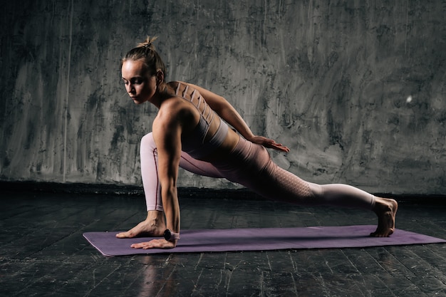 Mujer atlética joven muscular con hermoso cuerpo perfecto vistiendo ropa deportiva practicando postura en estera de yoga. Mujer caucásica de fitness posando en estudio con fondo gris oscuro.