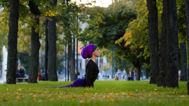 Mujer atlética musulmana practica el saludo al sol de yoga en el parque en la rutina matutina de hierba frente al perro