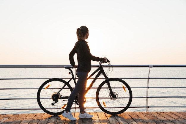 Mujer atlética de pie con bicicleta en el paseo marítimo, durante el amanecer sobre el mar