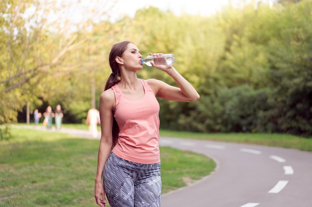 Mujer atlética de pie en la pista de atletismo en el parque de verano bebe agua después de los ejercicios de carrera Entrenamiento matutino femenino caucásico Jogging Concepto de estilo de vida saludable Entrenamiento cardiovascular tiro medio