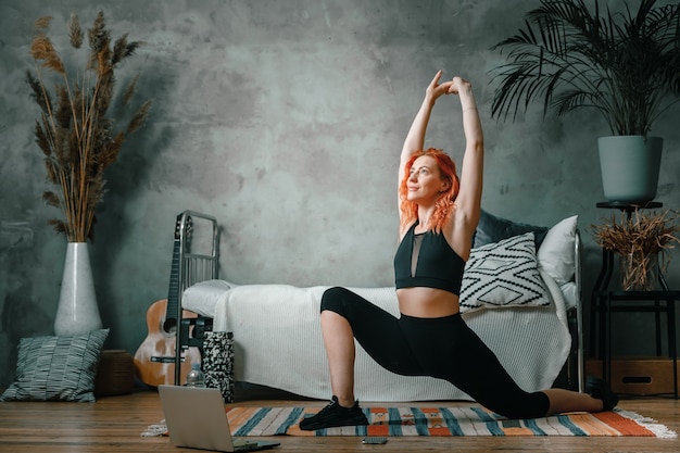 Mujer atlética positiva con cabello rojo y ropa deportiva ajustada se lanza en casa.