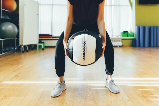 Mujer atlética que ejercita con la bola pesada grande mientras que está en la posición agazapada. Mujer muscular que hace entrenamiento cruzado del ajuste en el gimnasio.