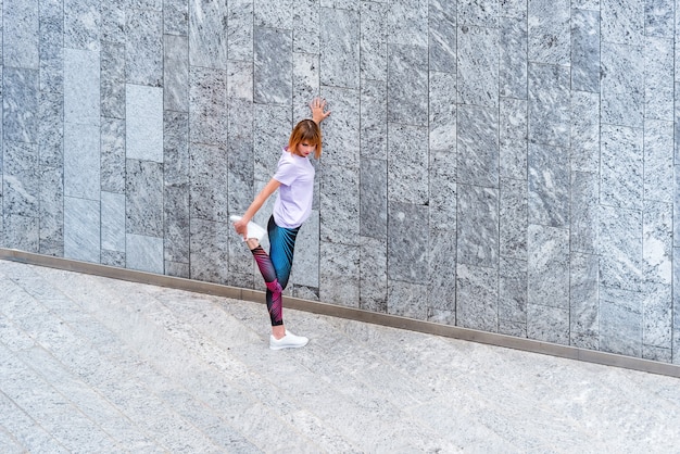 Mujer atlética que trabaja en la ciudad calentando sus músculos haciendo ejercicios de estiramiento de piernas en una pasarela junto a un edificio comercial en un concepto de salud y fitness