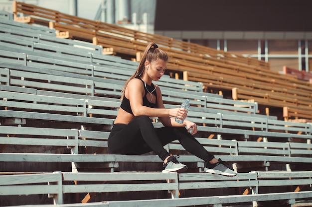 Mujer atlética en ropa deportiva y auriculares escuchando música y bebiendo agua de botella