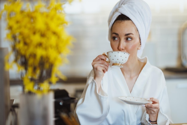 Una mujer atractiva en bata de baño bebiendo café por la mañana, disfrutando y relajándose en la cocina de su casa.