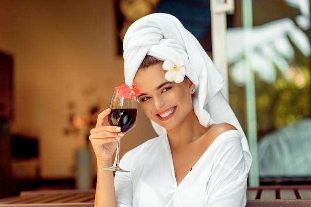 Mujer atractiva en una bata de baño blanca y una toalla con copa de vino y sonriendo a la cámara