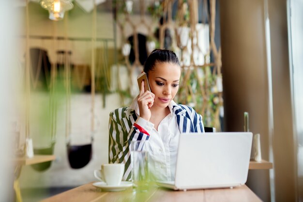 Mujer atractiva en un café, hablando por su teléfono y trabajando en equipo portátil.