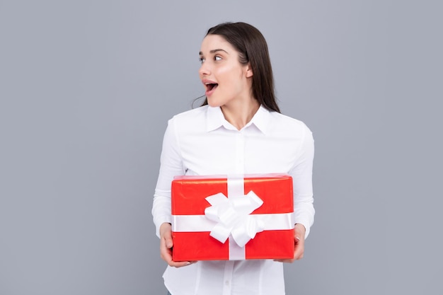 Mujer atractiva con caja presente posando con expresión de cara sorprendida sobre fondo gris Chica sosteniendo regalo decorado con cinta Día de San Valentín cumpleaños feliz año nuevo regalo personas concepto