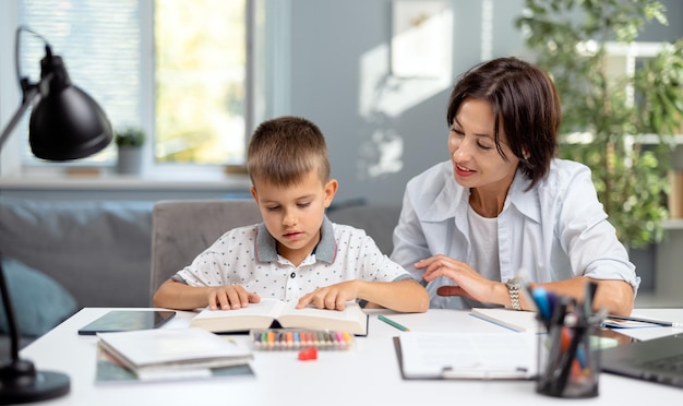 Mujer atractiva con camisa blanca sentada en la mesa con un niño pequeño y leyendo un libro juntos Madre caucásica cariñosa ayudando a su hijo con la tarea Concepto de familia y educación