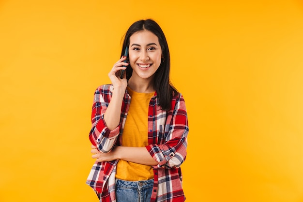 Mujer atractiva complacida en camisa a cuadros sonriendo y hablando celular aislado sobre pared amarilla