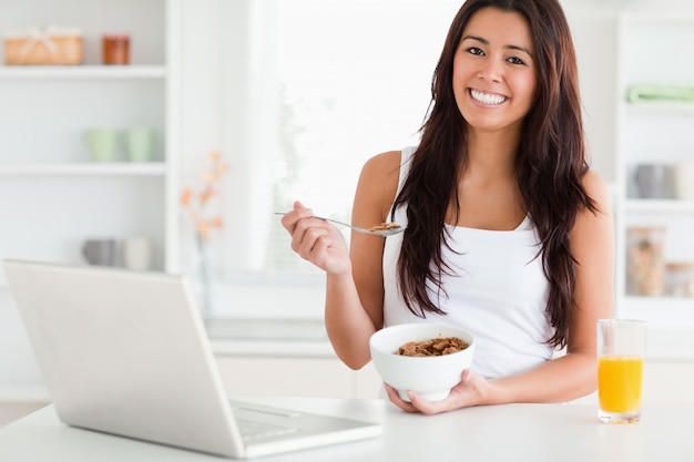 Mujer atractiva disfrutando de un tazón de cereales mientras se relaja con su computadora portátil