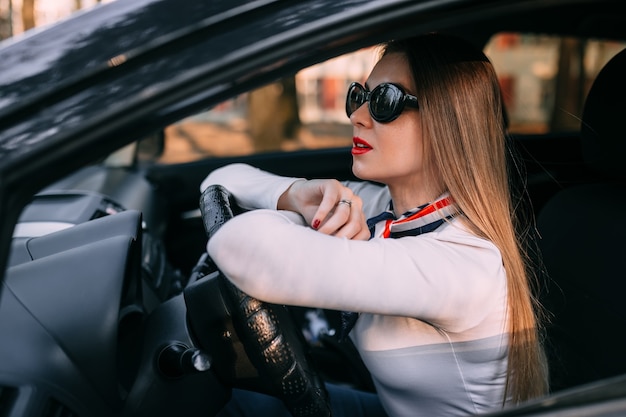 Mujer atractiva elegante con labios rojos en gafas de sol sentado en el coche y sosteniendo el volante