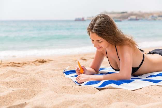 Mujer atractiva está acostada sobre una toalla rayada en la playa y exprimiendo bloqueador solar de un tubo en su mano.