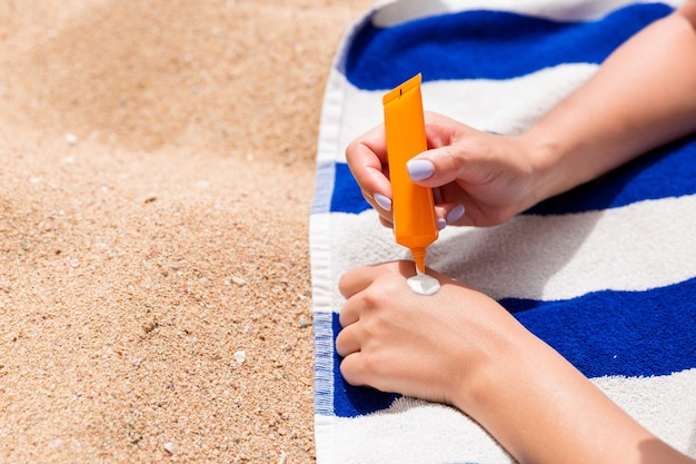 Mujer atractiva está acostada sobre una toalla rayada en la playa y exprimiendo bloqueador solar de un tubo en su mano.