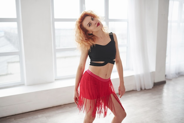 Mujer atractiva en falda roja posando en la habitación espaciosa cerca de la ventana.