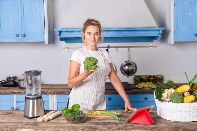 Mujer atractiva feliz en delantal sosteniendo brócoli y sonriendo a la cámara, cocinando ensalada en la cocina moderna, preparando comida vegetariana, cesta de verduras frescas en la mesa, dieta vegana nutrición saludable