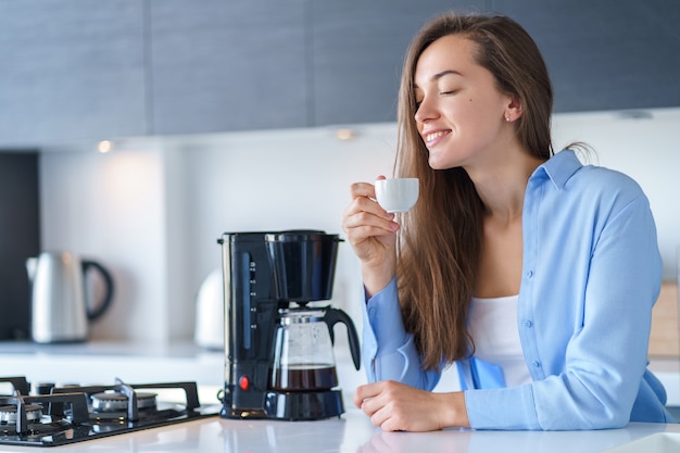 Mujer atractiva feliz que goza del aroma fresco del café después de preparar el café usando la cafetera en la cocina en casa. Batidora de café y electrodomésticos para hacer bebidas calientes