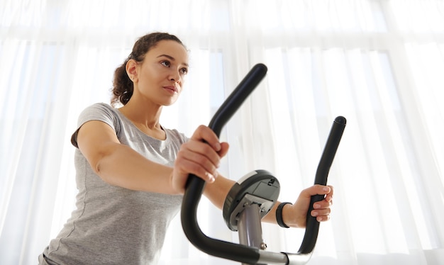 Mujer atractiva en forma de ejercicio en una bicicleta de spinning en casa en un hermoso día soleado