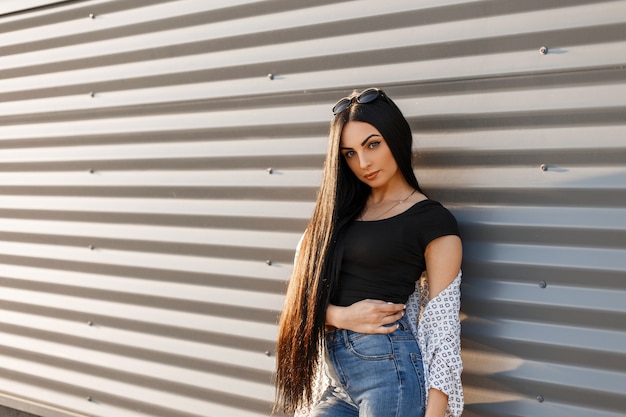 Mujer atractiva hermosa hipster en gafas redondas con estilo oscuro en ropa de verano con estilo posa cerca de una pared de metal moderno al atardecer