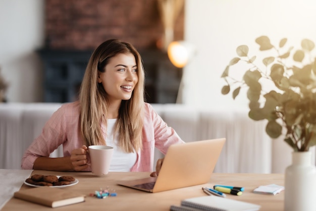 Mujer atractiva inspirada sonriendo y mirando hacia un lado mientras se sienta frente a la computadora portátil en una habitación iluminada por el sol