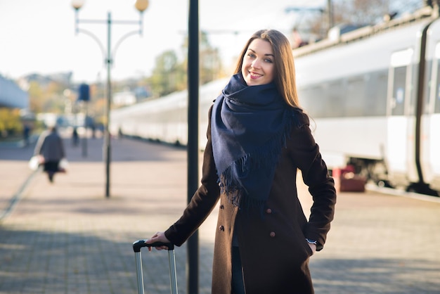 Mujer atractiva joven en abrigo con una maleta esperando en la estación de tren