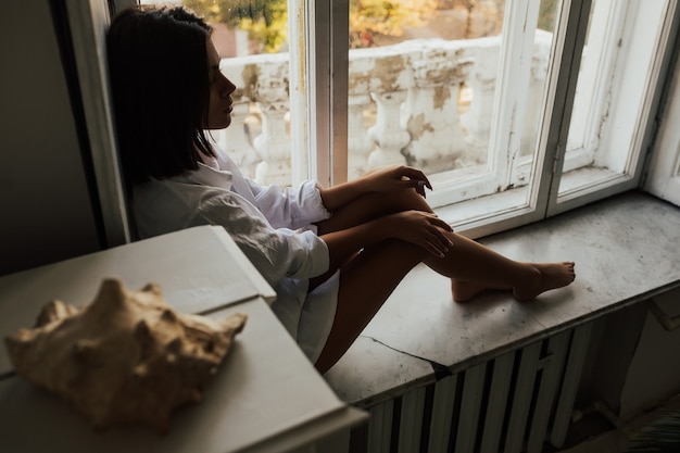 Mujer atractiva joven en camisa blanca masculina, sentado en un alféizar de la ventana, estado de ánimo solitario