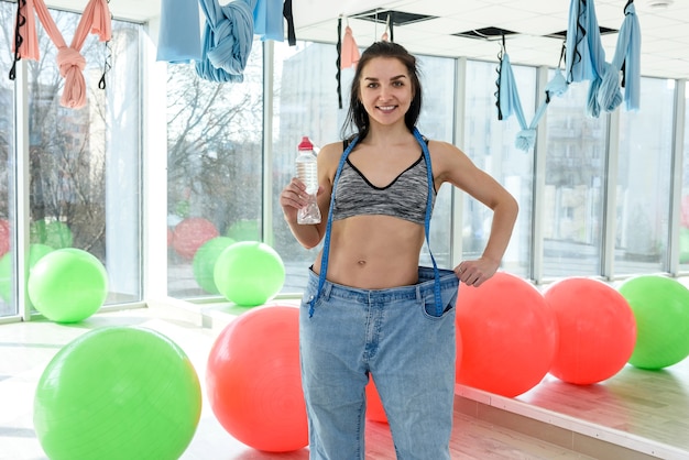 Mujer atractiva joven delgada con botella de agua y muestra el resultado de la pérdida de peso en el gimnasio o gimnasio. Estilo de vida saludable