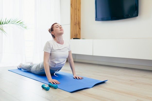 Mujer atractiva joven está practicando yoga en línea mediante una aplicación de teléfono móvil o teléfono inteligente. Mañana yogui femenina en ropa casual en casa en la sala de estar, posición de loto de entrenamiento medita sobre una estera