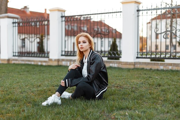 Mujer atractiva joven estadounidense con una elegante chaqueta en jeans negros con zapatillas blancas se sienta en un césped verde de un edificio blanco vintage al atardecer. Niña bonita.