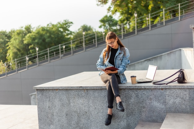 La mujer atractiva joven del estudiante está leyendo un libro mientras que se sienta en las escaleras en la ciudad. El concepto de aprendizaje a distancia. Estudiante moderno