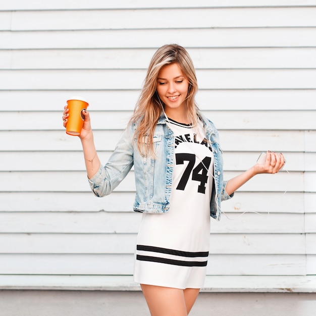 Mujer atractiva joven feliz en ropa de verano de moda posando junto a la pared de madera vintage en un día cálido y soleado. Encantadora chica bebe café y disfruta del fin de semana.