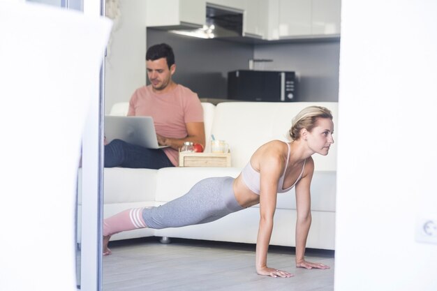 Mujer atractiva joven haciendo ejercicios de fitness y hombre trabajando en el sofá con una computadora