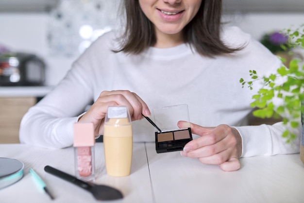 Mujer atractiva joven haciendo maquillaje, mujer sentada en su casa a la mesa en la cocina con pincel de espejo de productos cosméticos, muestra maquillaje de sombra de cejas. Belleza, cuidado de la piel, moda, concepto de maquillaje