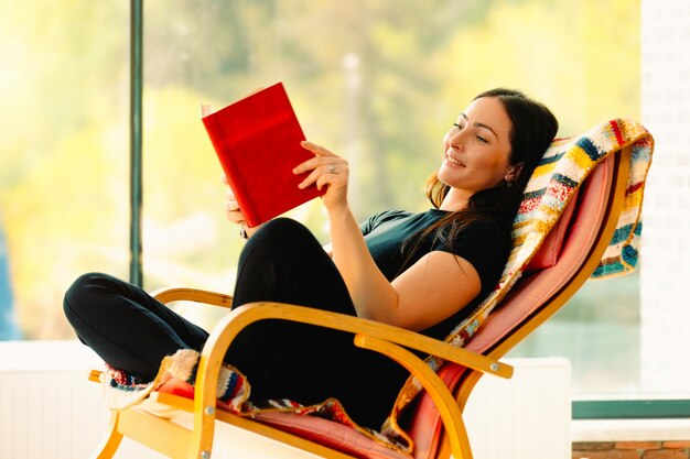 Foto mujer atractiva joven lee un libro meciéndose en una cómoda mecedora. .