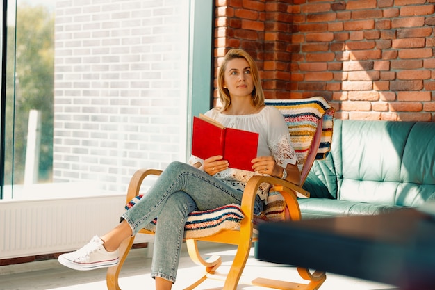 Mujer atractiva joven mira pensativamente a un lado con un libro en sus manos, balanceándose en una cómoda mecedora. Pasar un buen rato mejora tu estado de ánimo.