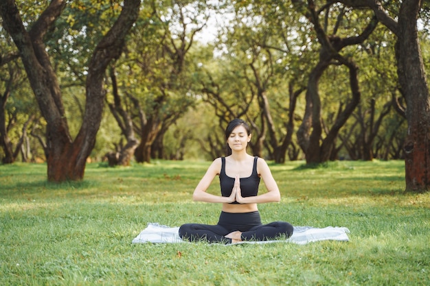 Foto mujer atractiva joven con los ojos cerrados meditando en posición de loto sentado en el césped en el parque.